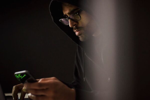 Crop bearded ethnic male computer geek in hoodie chatting on cellphone while typing on netbook in dark room
