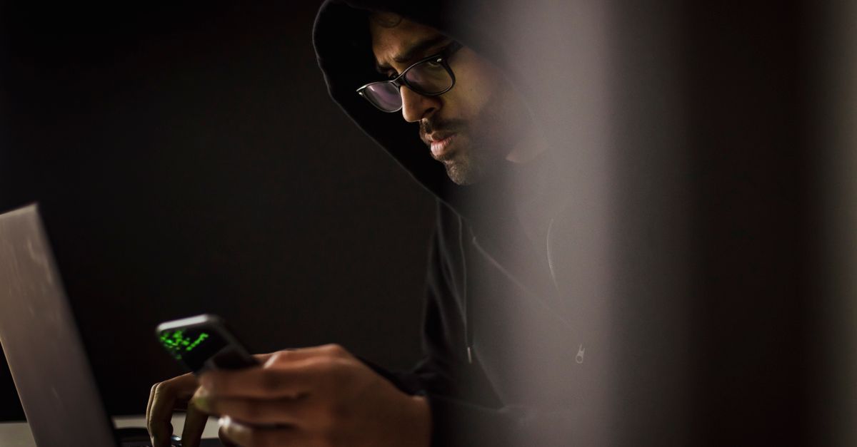 Crop bearded ethnic male computer geek in hoodie chatting on cellphone while typing on netbook in dark room