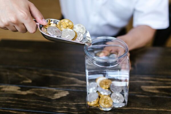 Hand placing gold and silver coins into a glass jar on a wooden table indoors.