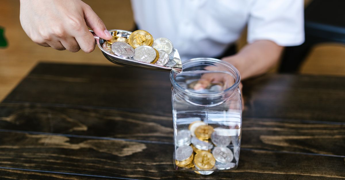 Hand placing gold and silver coins into a glass jar on a wooden table indoors.