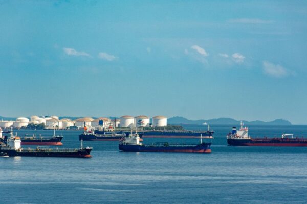 A fleet of cargo ships docked near oil storage tanks along a serene coastline with a clear blue sky above.