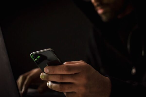Crop anonymous ethnic male cyber spy with cellphone and netbook hacking system in evening