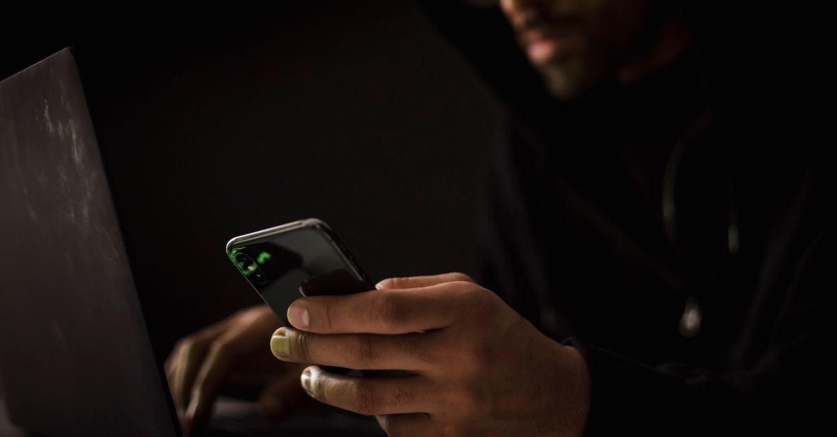 Crop anonymous ethnic male cyber spy with cellphone and netbook hacking system in evening