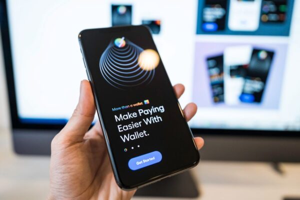 Hand holding smartphone displaying digital wallet app interface, blurred monitor in background.